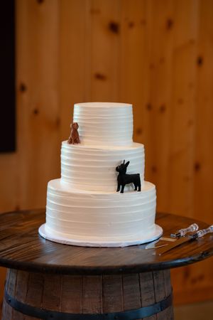 Three-tiered white wedding cake decorated with black silhouettes of animals sits atop wooden barrel.