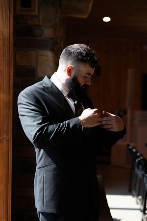 Man in dark suit adjusts his tie while standing in dramatic lighting.