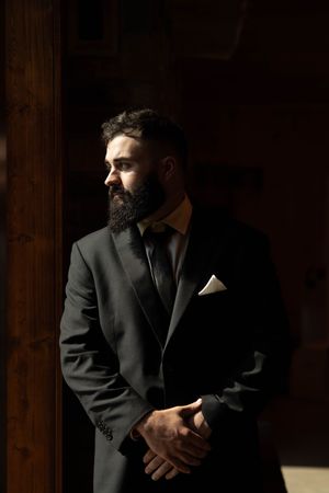 Dramatic portrait of a bearded man in formal suit standing by window in moody lighting.
