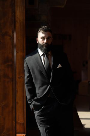 Portrait of suited man with full beard standing against wooden wall in dramatic lighting.