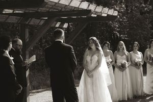 Wedding ceremony setup with white chairs and bridal party under wooden pergola.