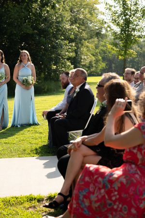Wedding guests watching ceremony from white chairs on sunny lawn.