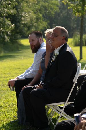 Wedding guests seated on white chairs during outdoor ceremony in sunlit garden.