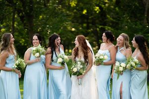 Bridesmaids in light blue dresses stand together holding bouquets outdoors.
