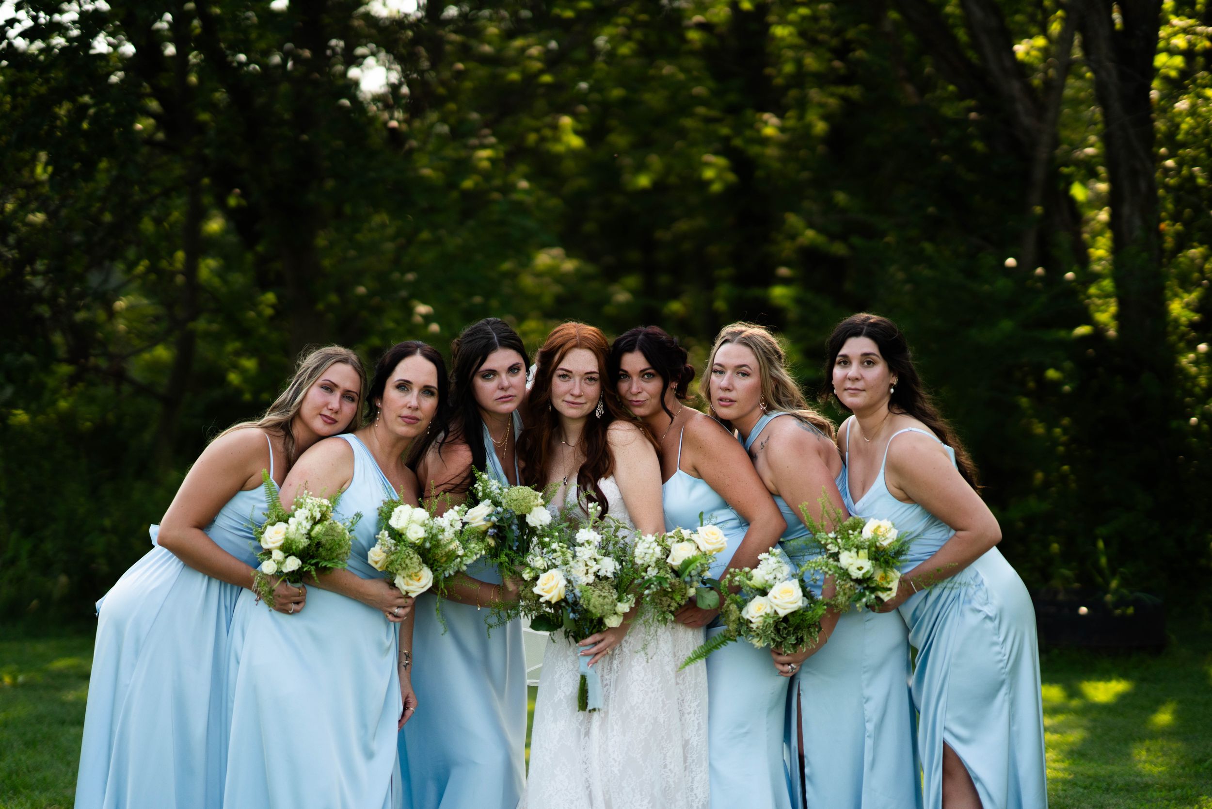 Bridesmaids in light blue dresses pose together outdoors holding white floral bouquets.