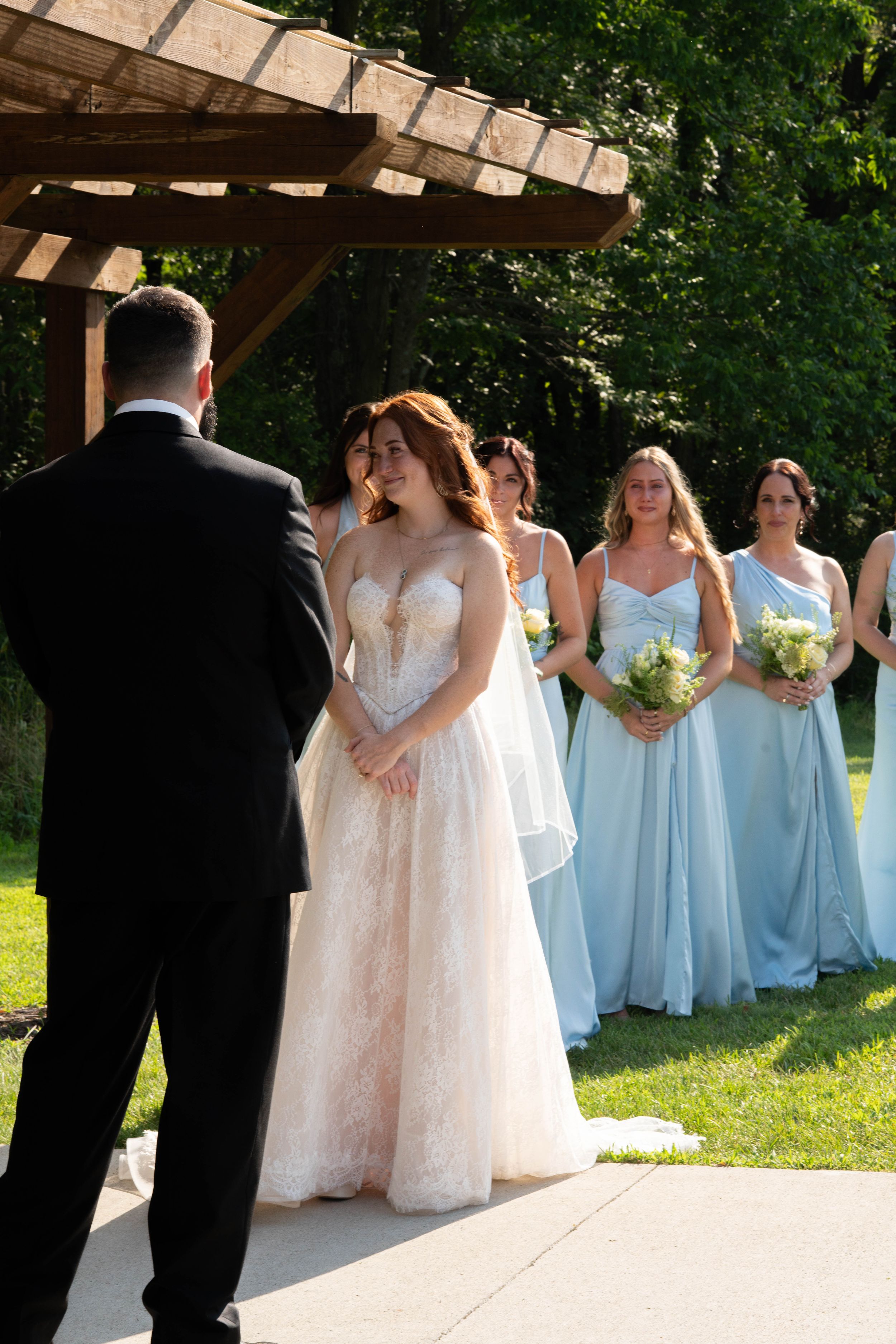 Wedding party in light blue dresses and formal attire poses under a wooden pergola.