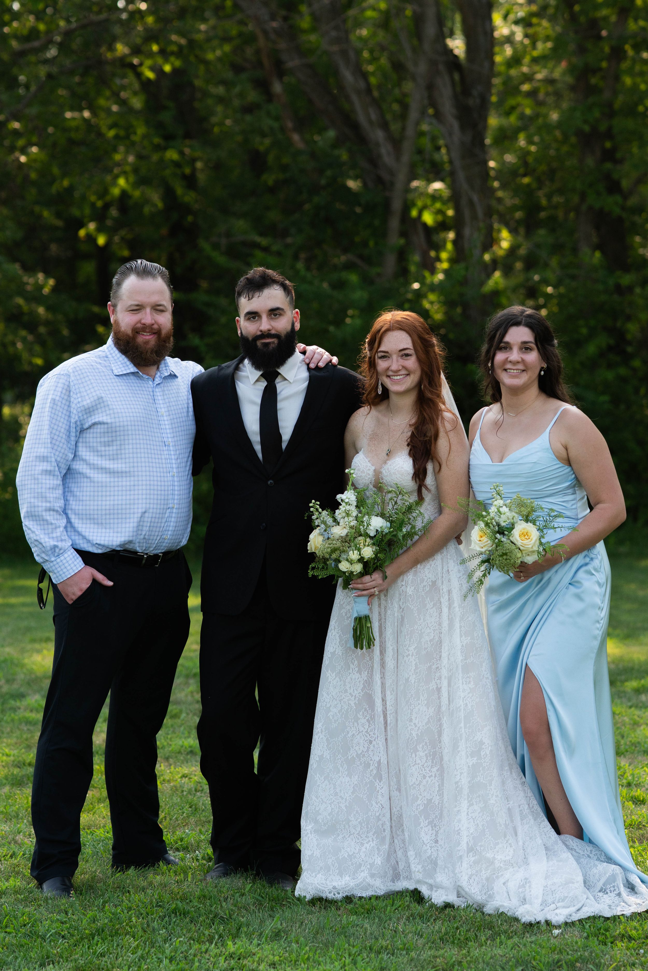Wedding party poses together on grassy lawn with trees in background wearing formal attire and light blue dresses.