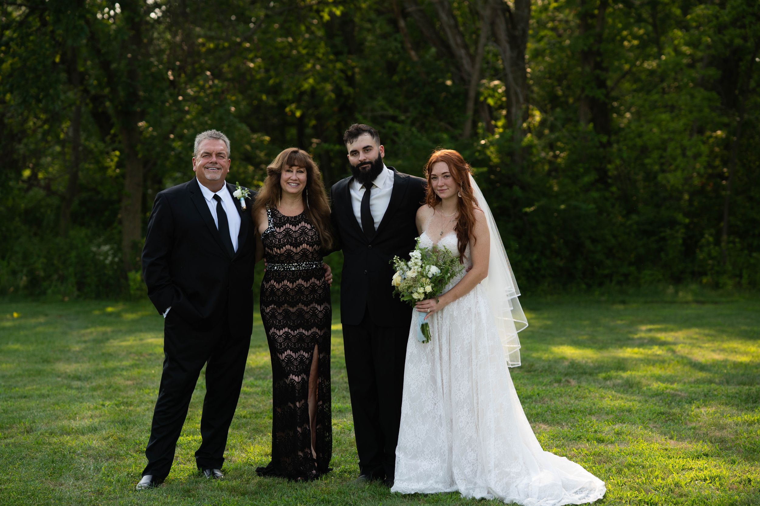 Wedding party members stand together outdoors in formal wear against a natural green backdrop.