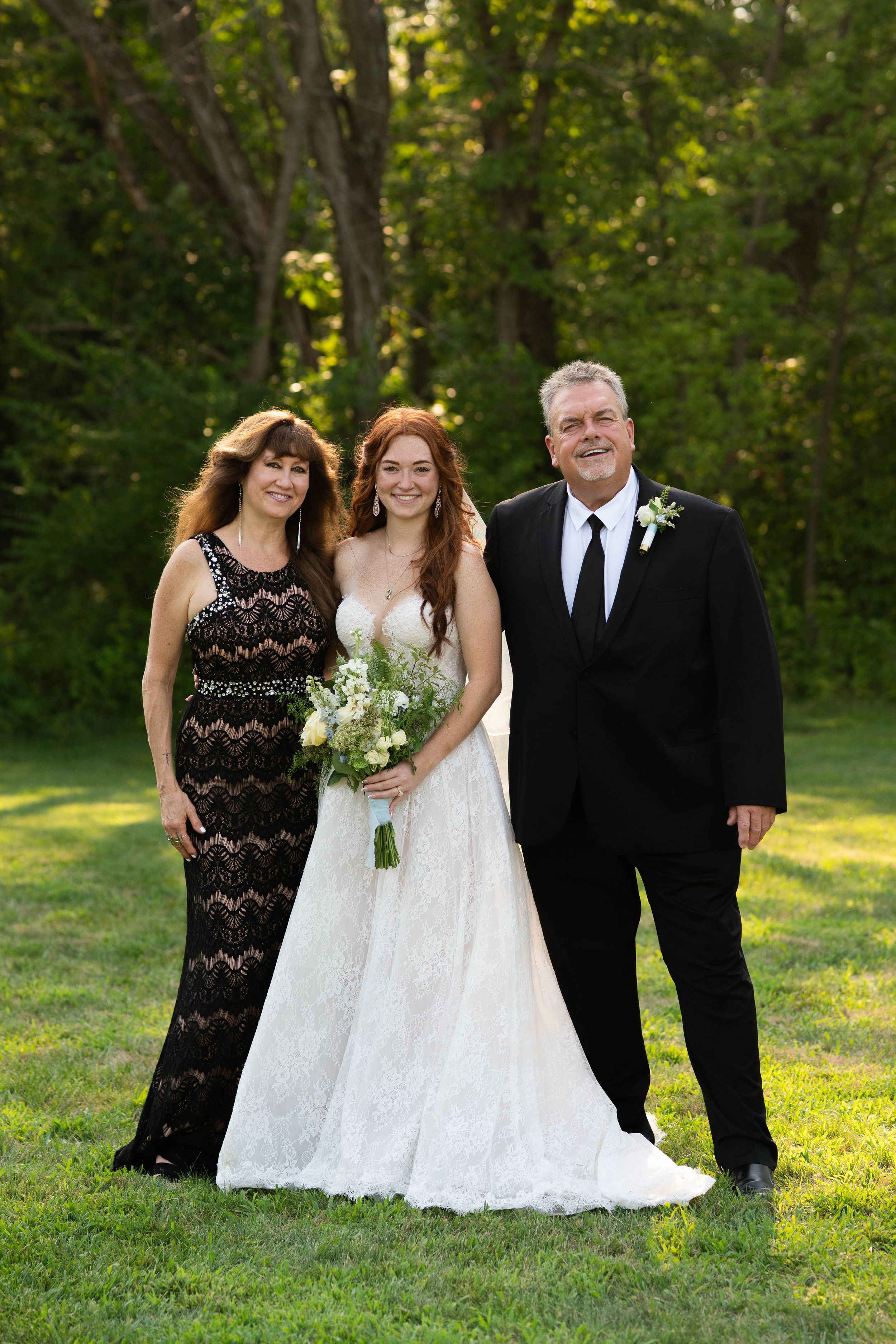 Wedding party members gather for formal photos on grass with trees in background during golden hour.