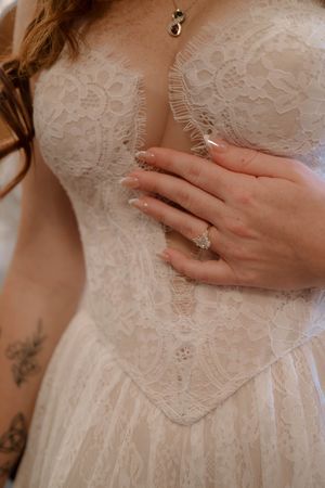 Detailed shot of intricate lace wedding dress bodice with engagement ring and small tattoo visible.