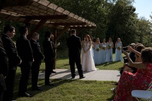 Outdoor wedding ceremony setup with white chairs and guests gathered under trees.