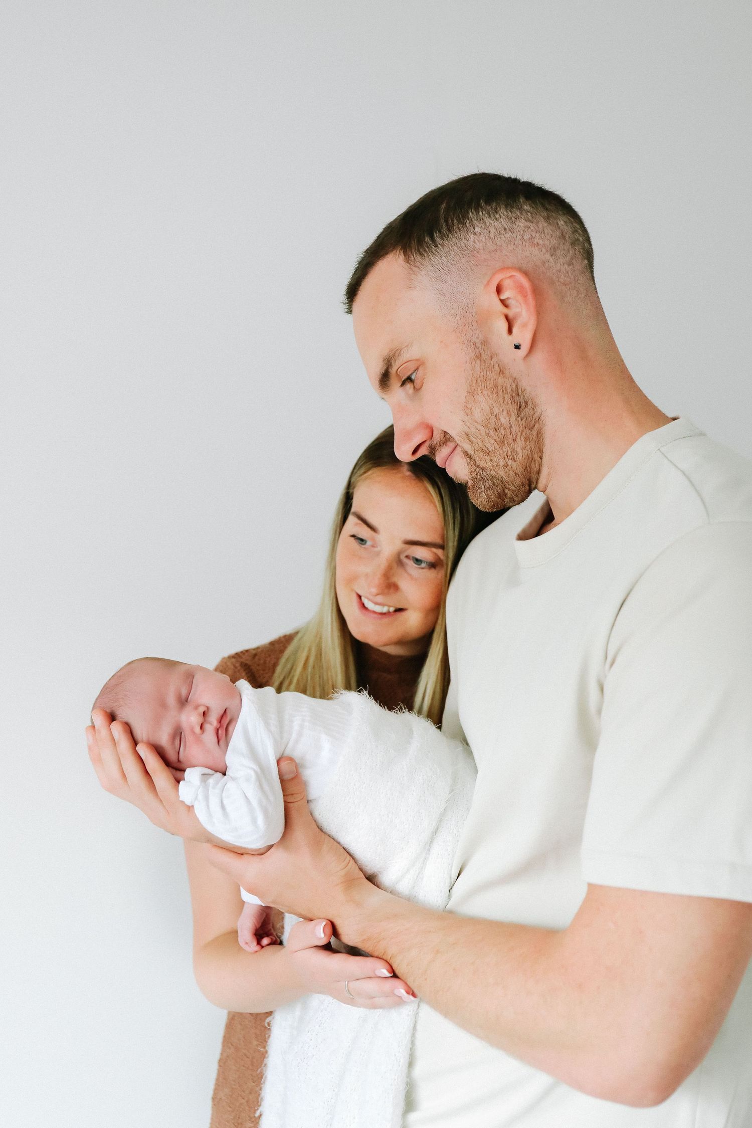 A couple in white holding their newborn baby wrapped in a blanket against a light background, during newborn shoot based in Nottingham