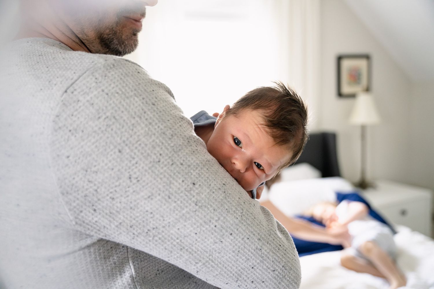 A small child peeks over a parent's shoulder while being held in a cozy bedroom setting.