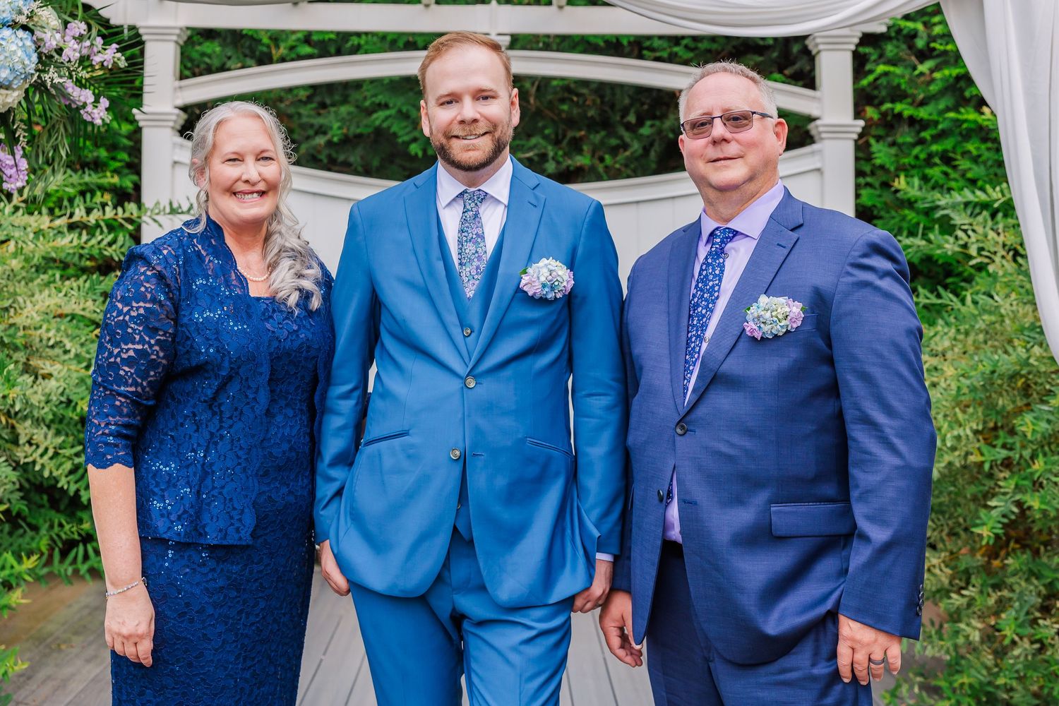 Wedding party gathered under a white canopy in an outdoor garden setting wearing navy and blue formal attire.