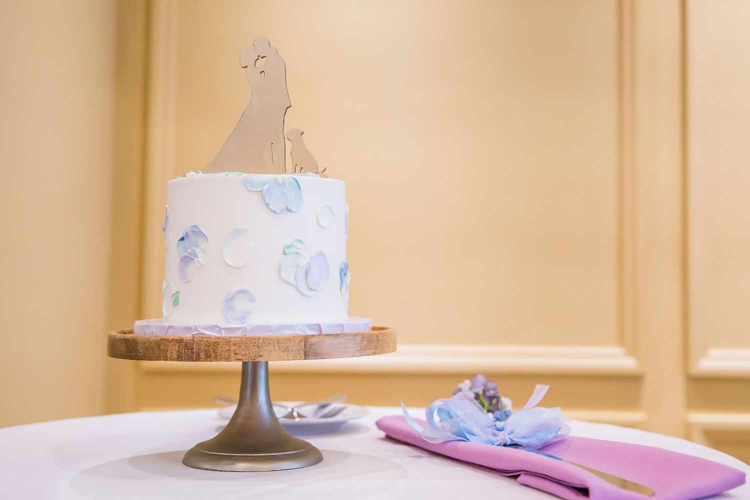 A white buttercream cake with blue designs sits on a pedestal stand next to a folded pink napkin.