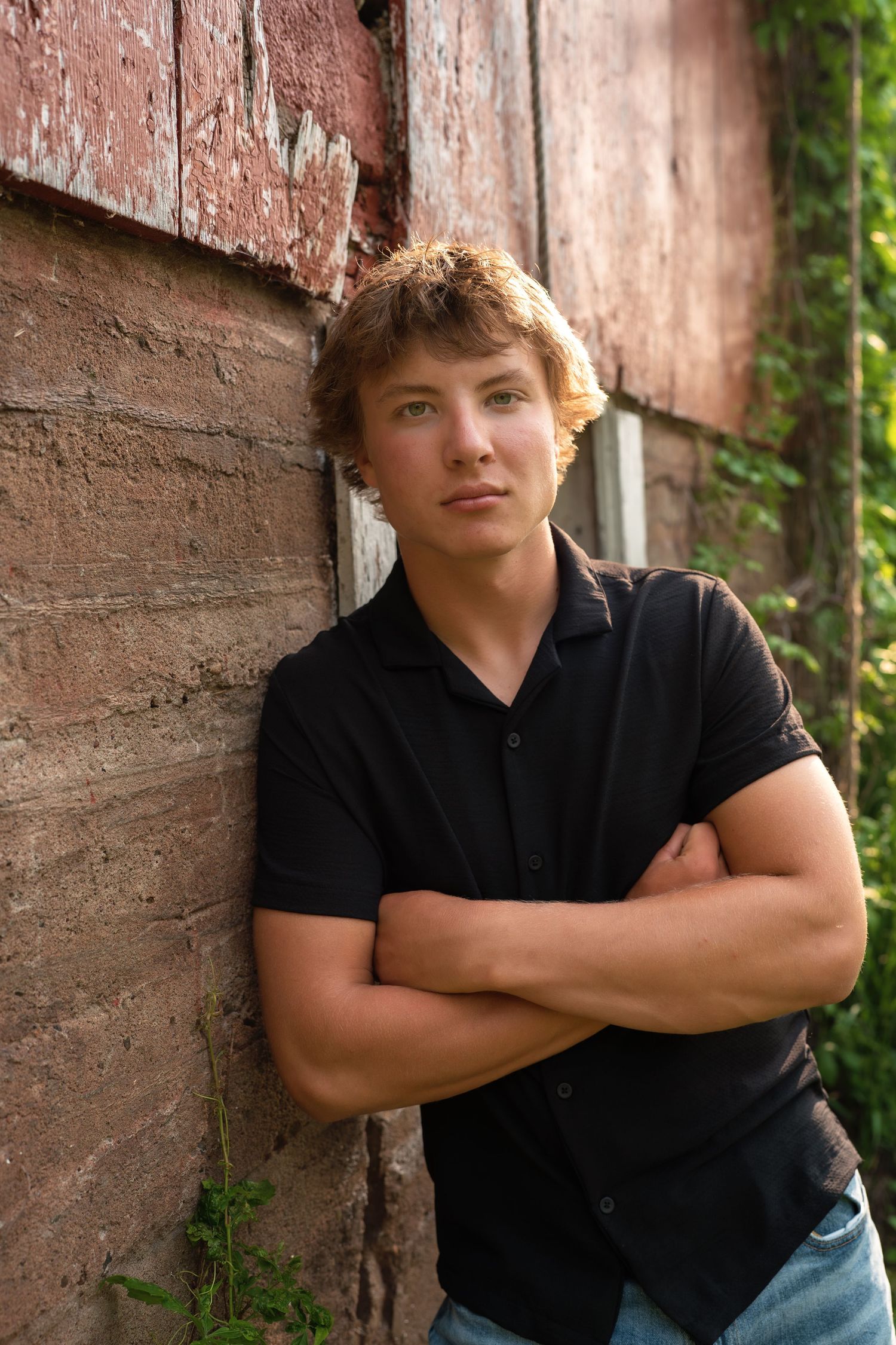 Casual outdoor portrait against rustic red wall with green foliage in background.