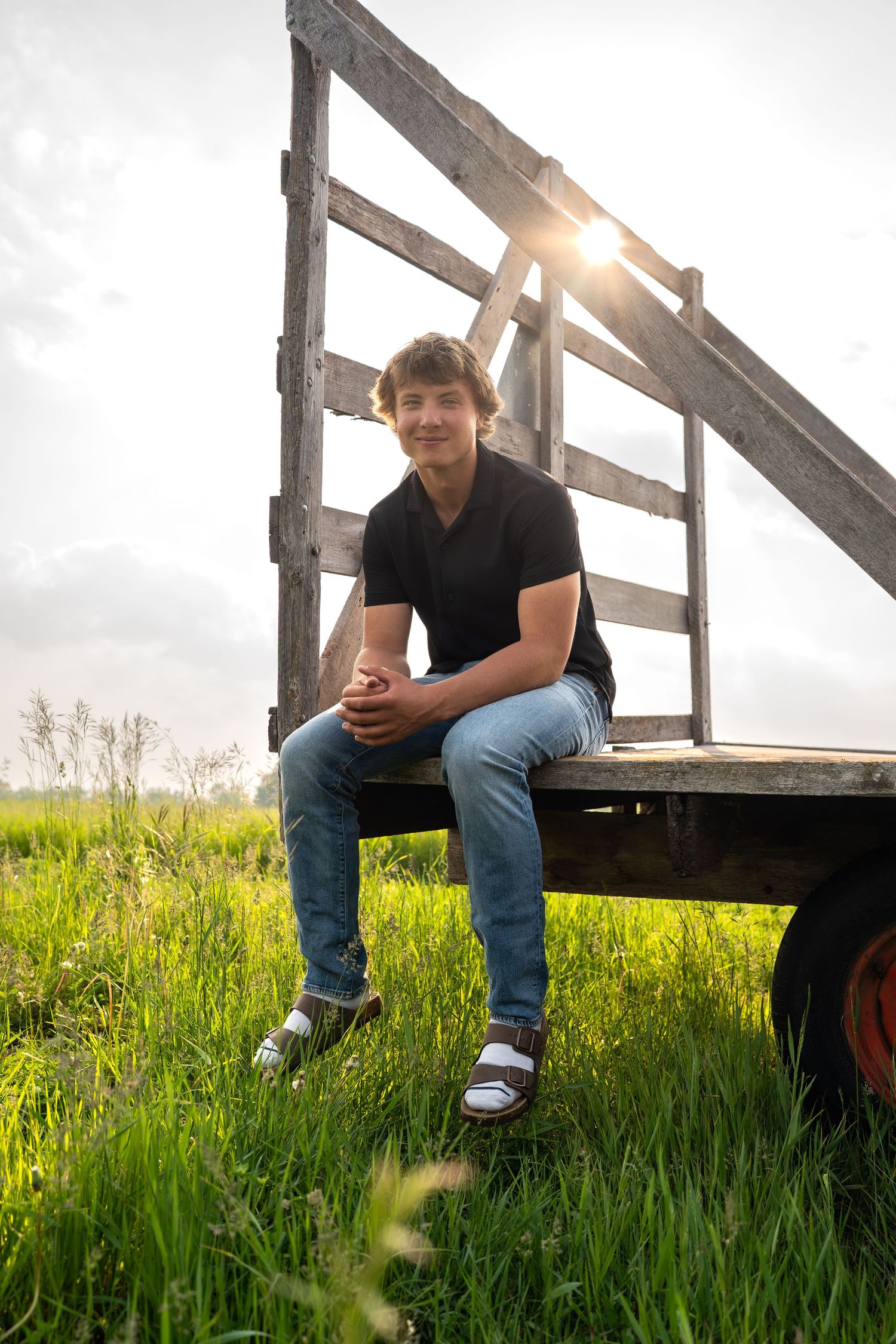 Person sits on wooden fence at sunset surrounded by tall grass in rural outdoor setting.