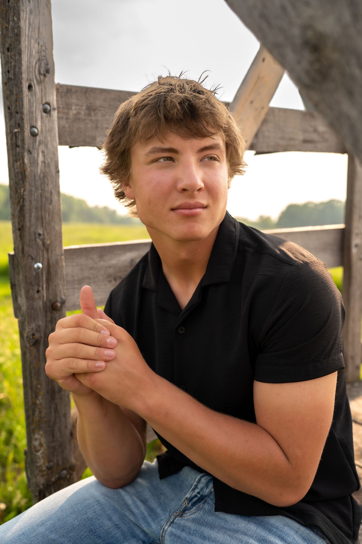 Young person in black polo shirt sitting outdoors against wooden fence post at sunset in rural setting.