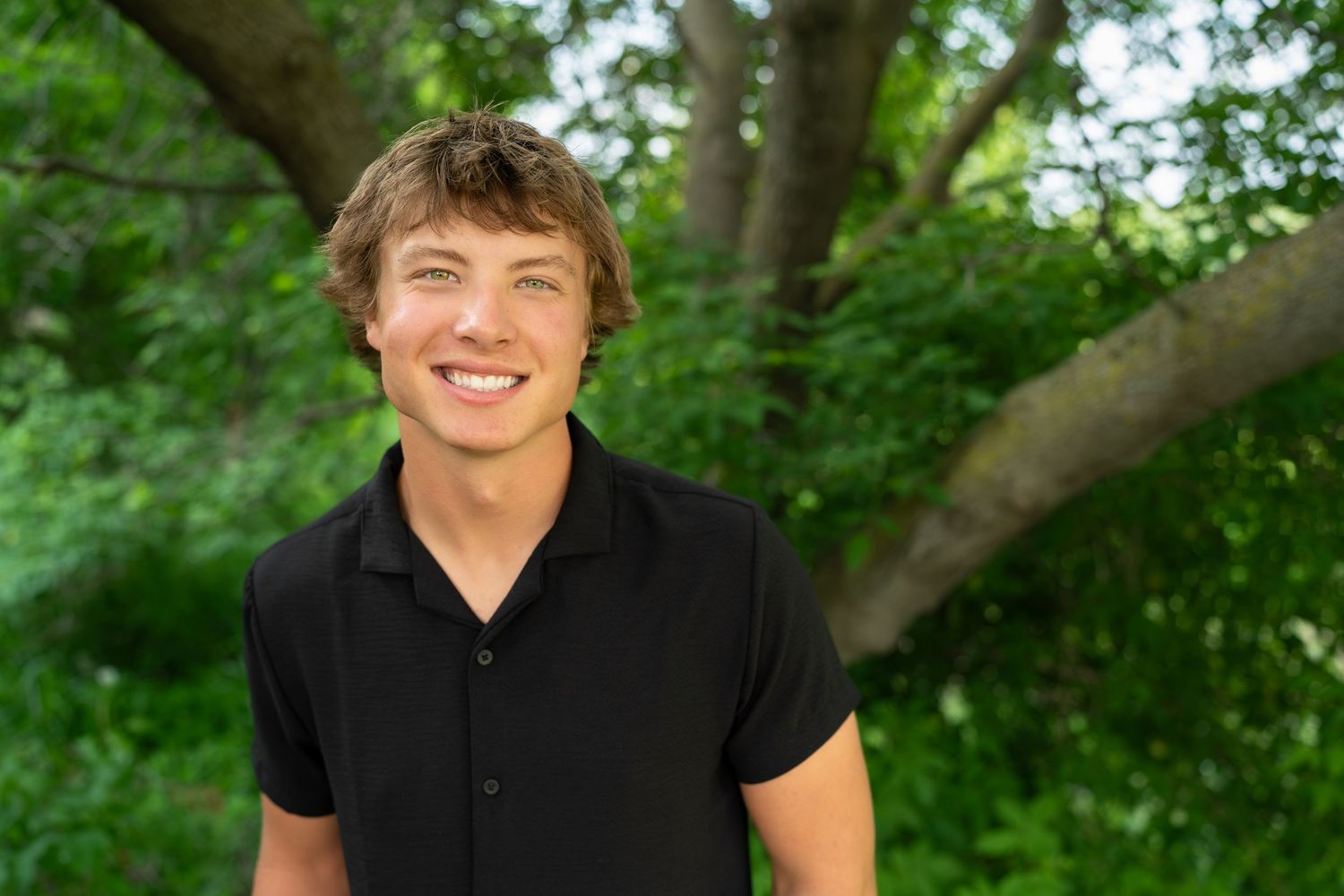 A bright smiling portrait of someone in a black polo shirt outdoors with green trees in the background.
