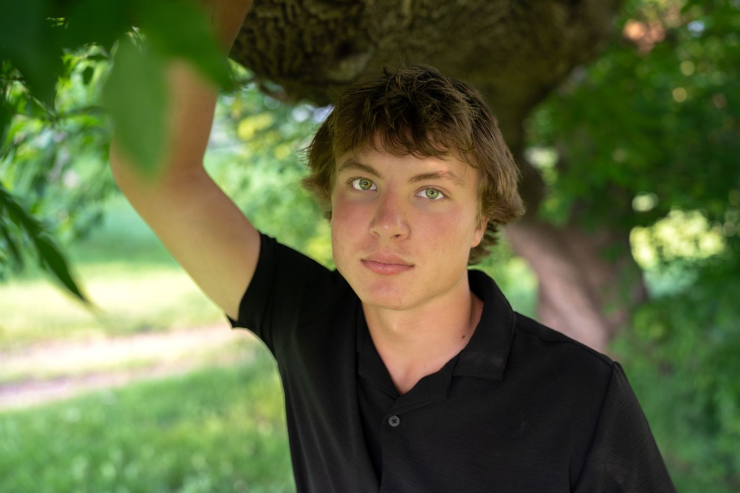 Person in black polo shirt standing under a tree in natural outdoor setting with green foliage in background.