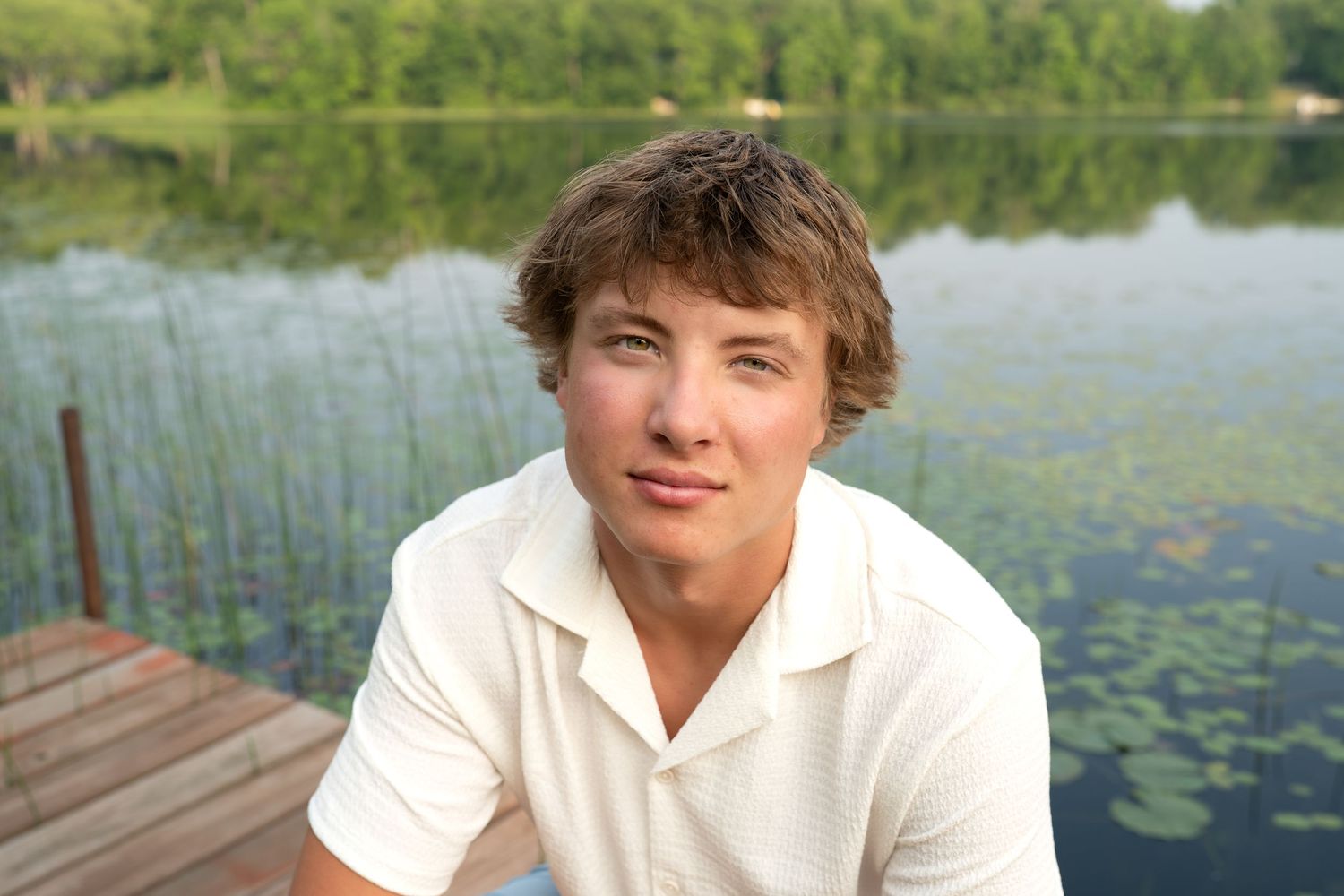 Person sitting on dock by serene lake with lily pads and lush green trees in background during summer.