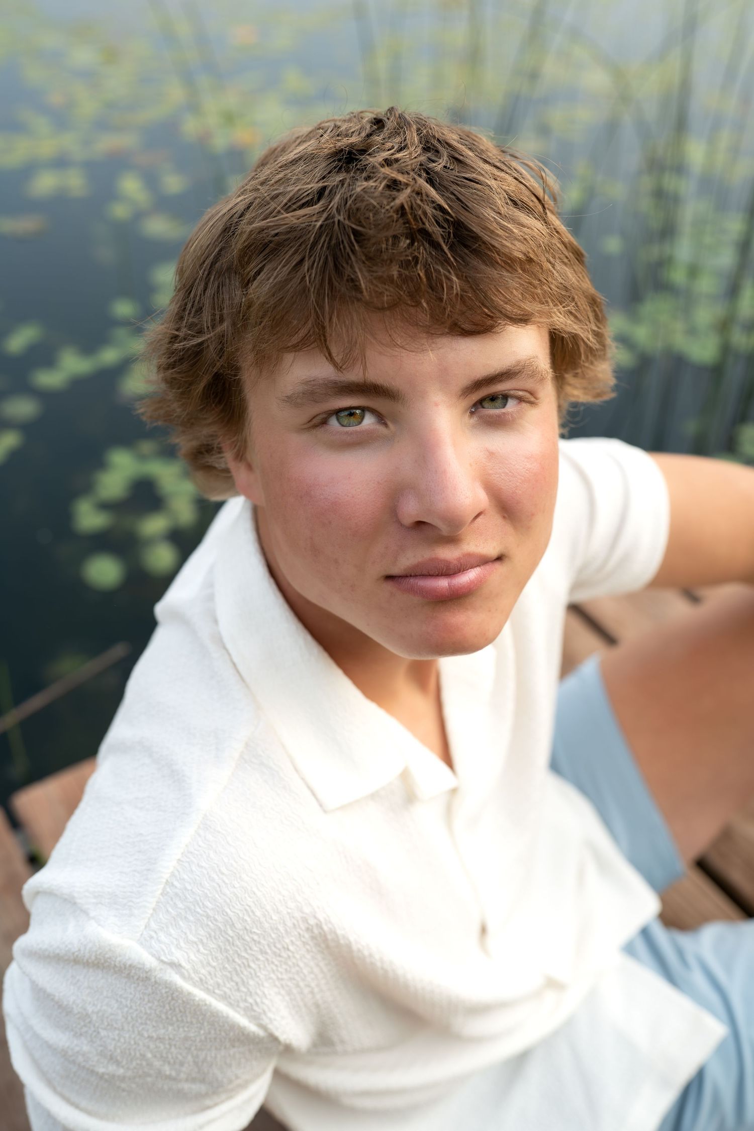 A person in a white sweater sits outdoors near water with lily pads visible in the background.