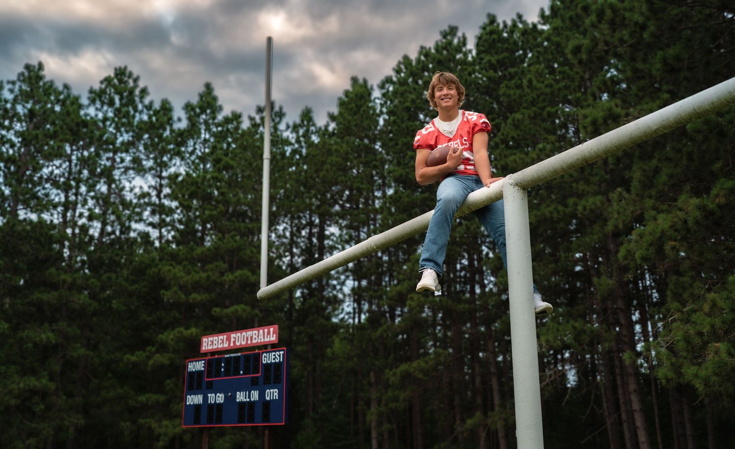 A person in an orange shirt sits atop a football goal post at sunset with pine trees and a scoreboard in the background.