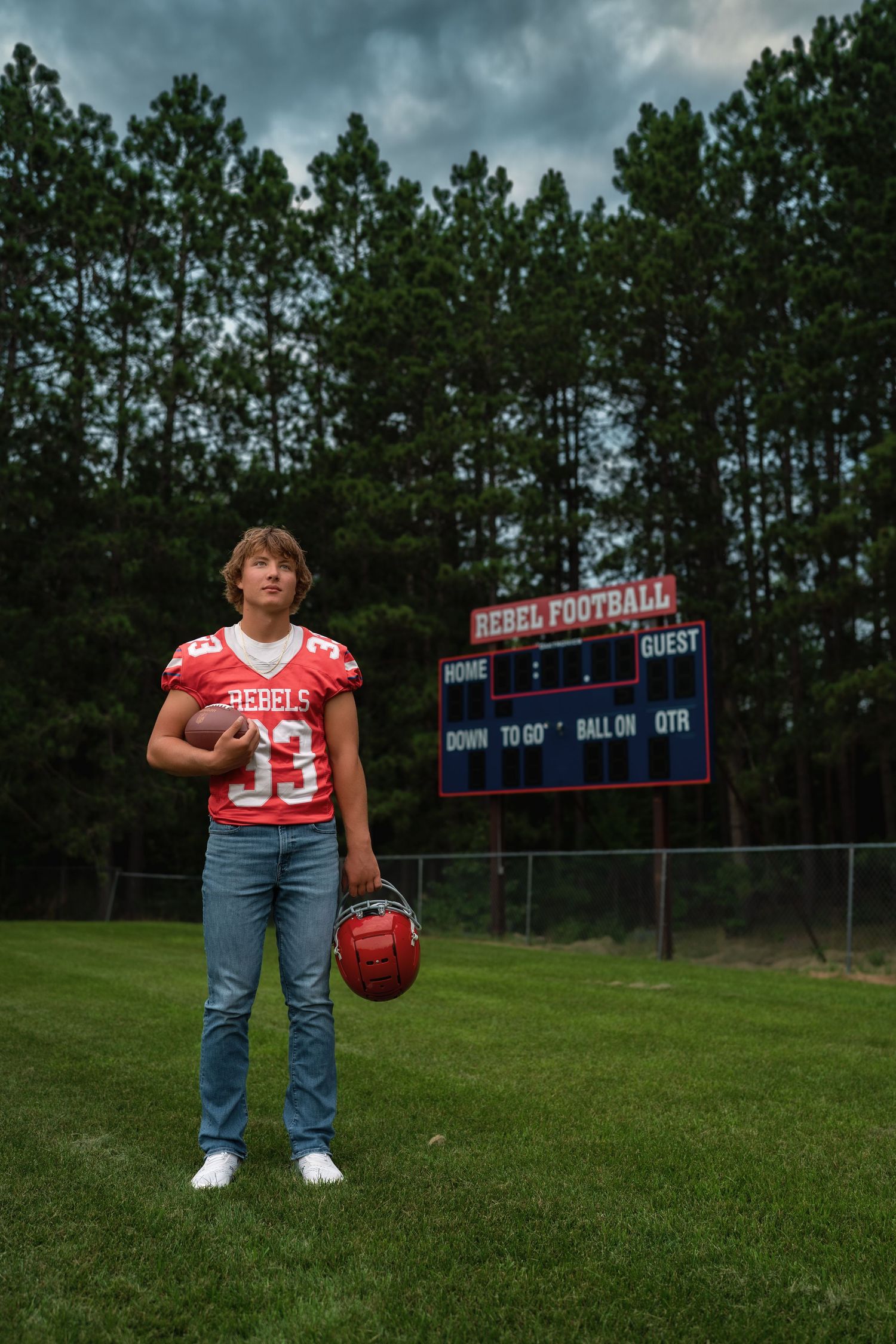 High school football player poses on field with scoreboard and storm clouds in background.