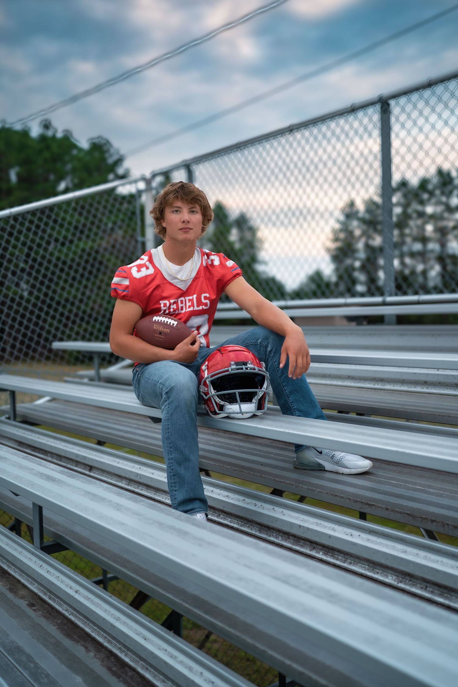 Football player sits on metal bleachers at sunset wearing red jersey and holding helmet in outdoor stadium setting.
