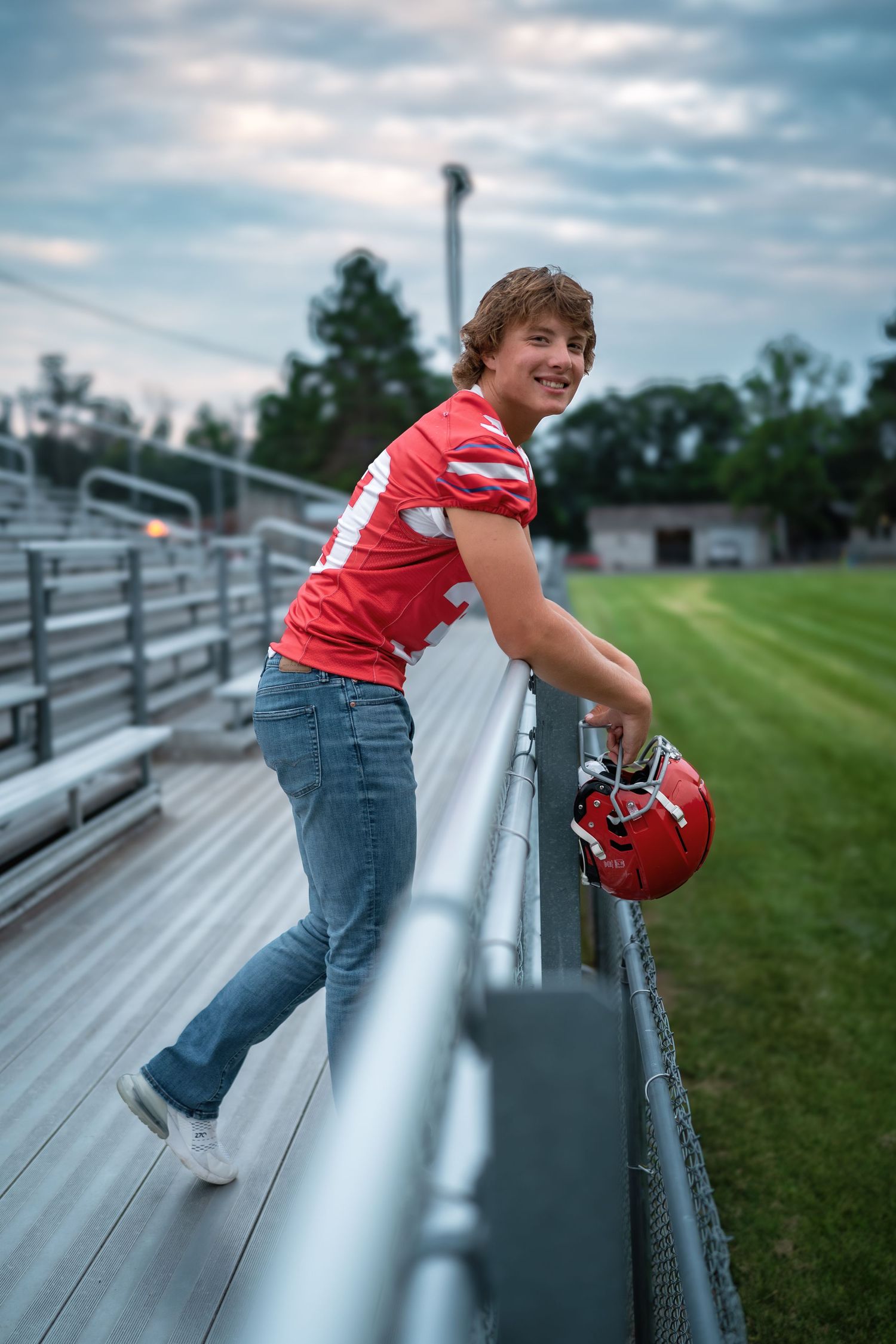 Student athlete in red jersey and jeans poses on bleachers with football helmet at empty stadium field.