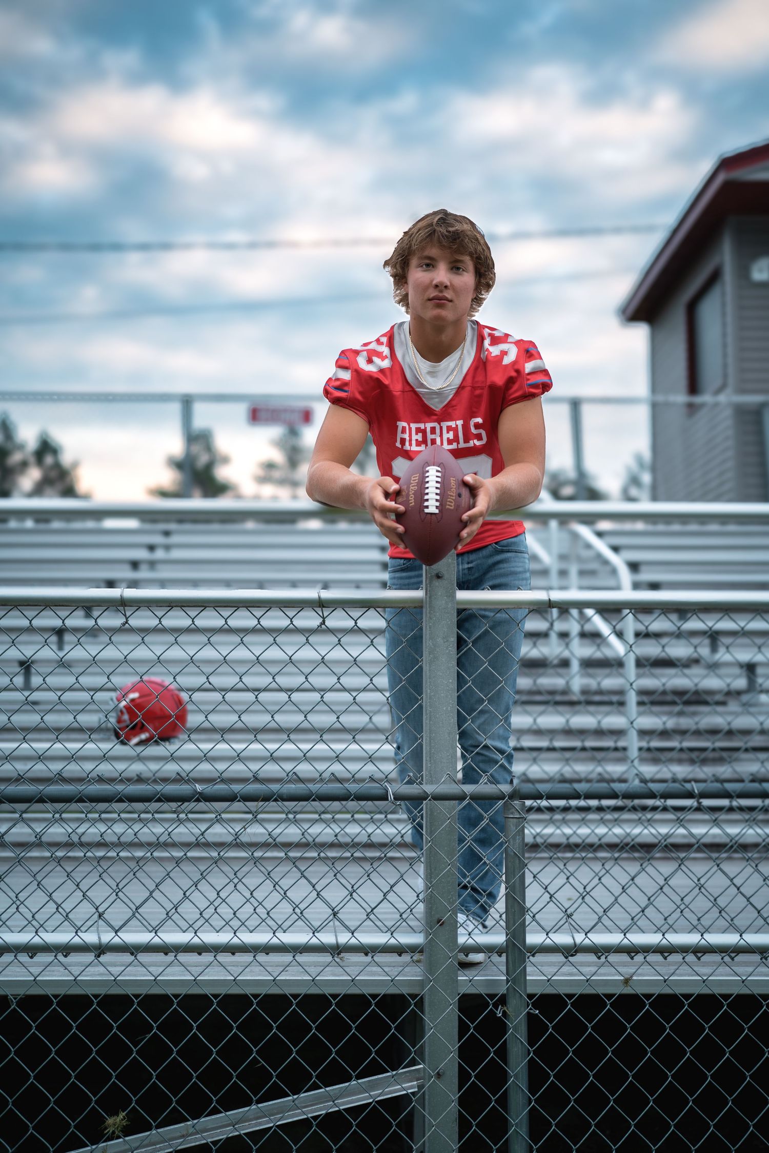 Football player in Rebels jersey leans on stadium railing with dramatic cloudy sky behind.