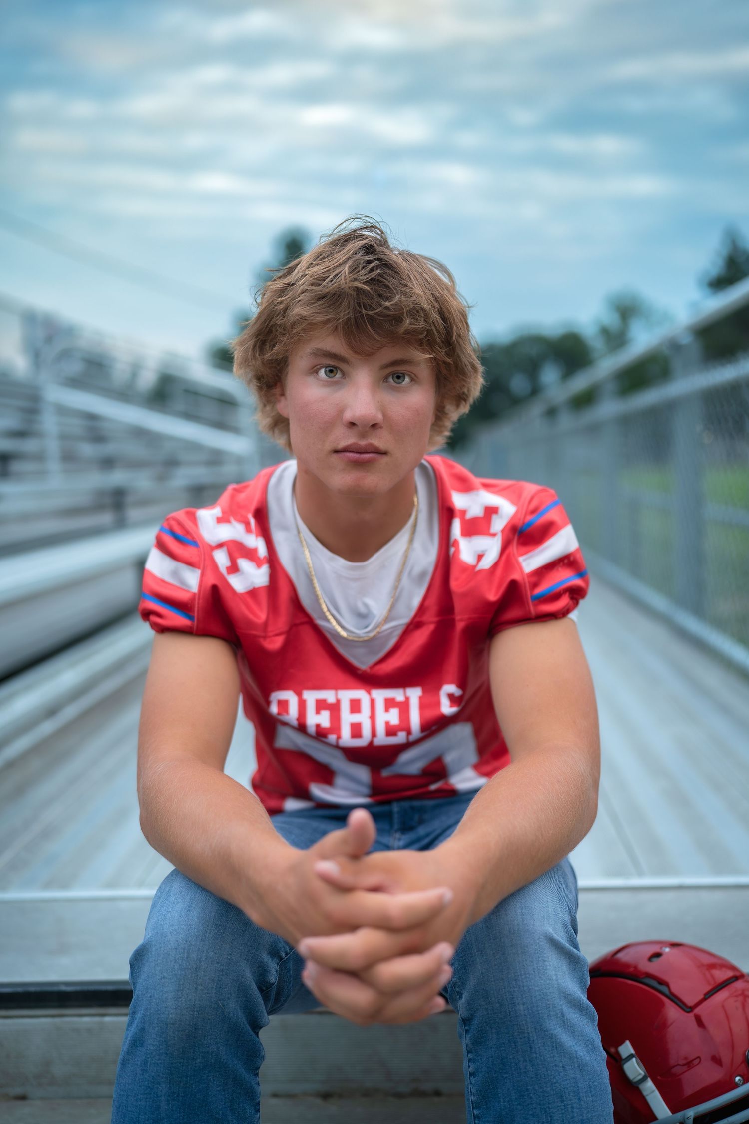 High school athlete in red and white Rebels football jersey sits thoughtfully on bleachers at sports field.