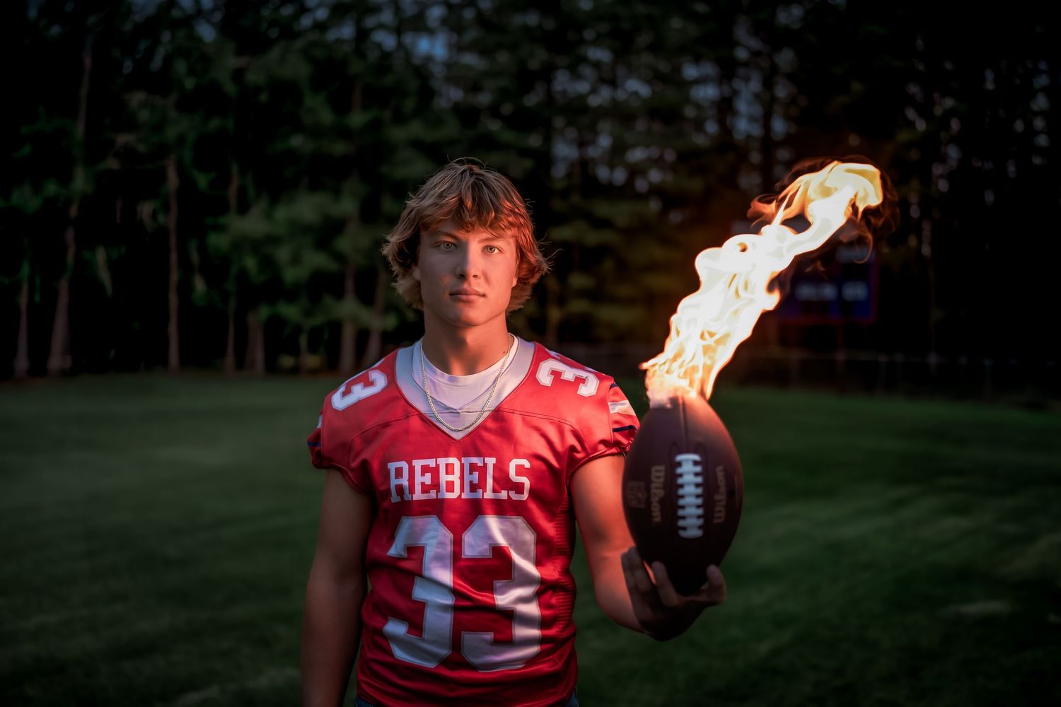 High school football player in red jersey poses with flaming football at twilight for dramatic sports portrait.