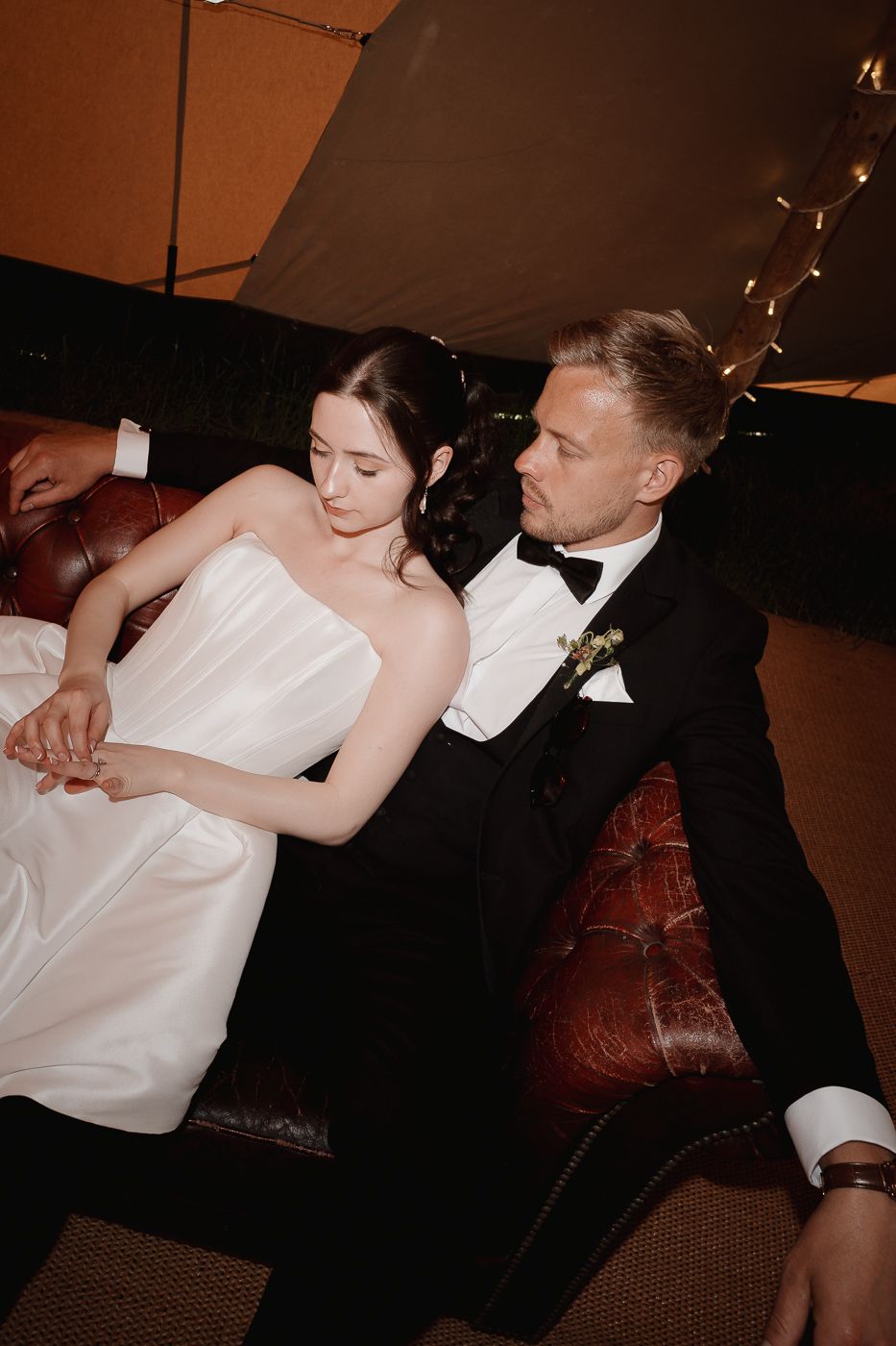 A romantic couple in formal wedding attire share intimate moments on a brown leather sofa under warm lighting.