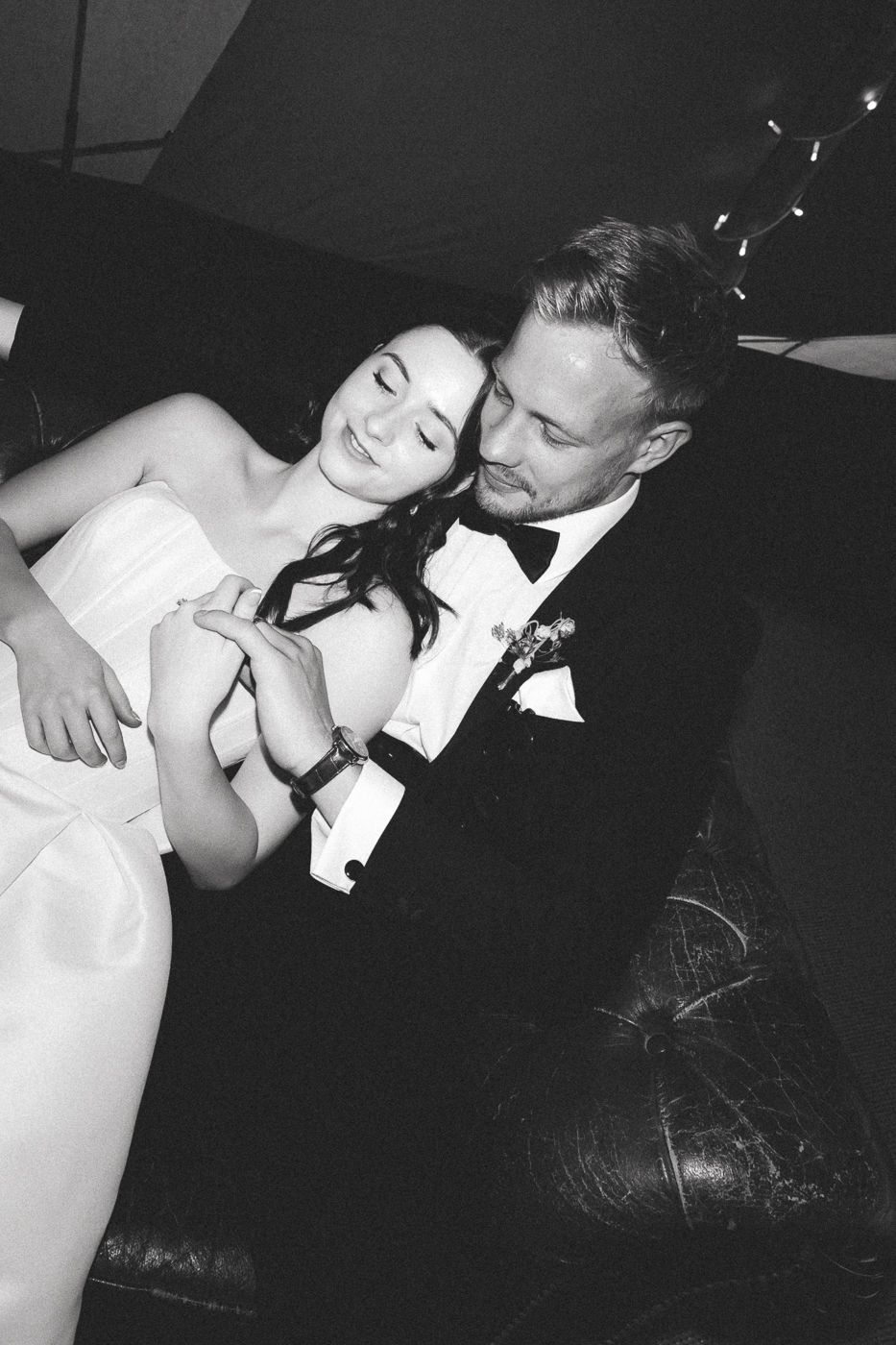 A black and white intimate wedding photo showing a couple sharing a tender moment on a leather couch.