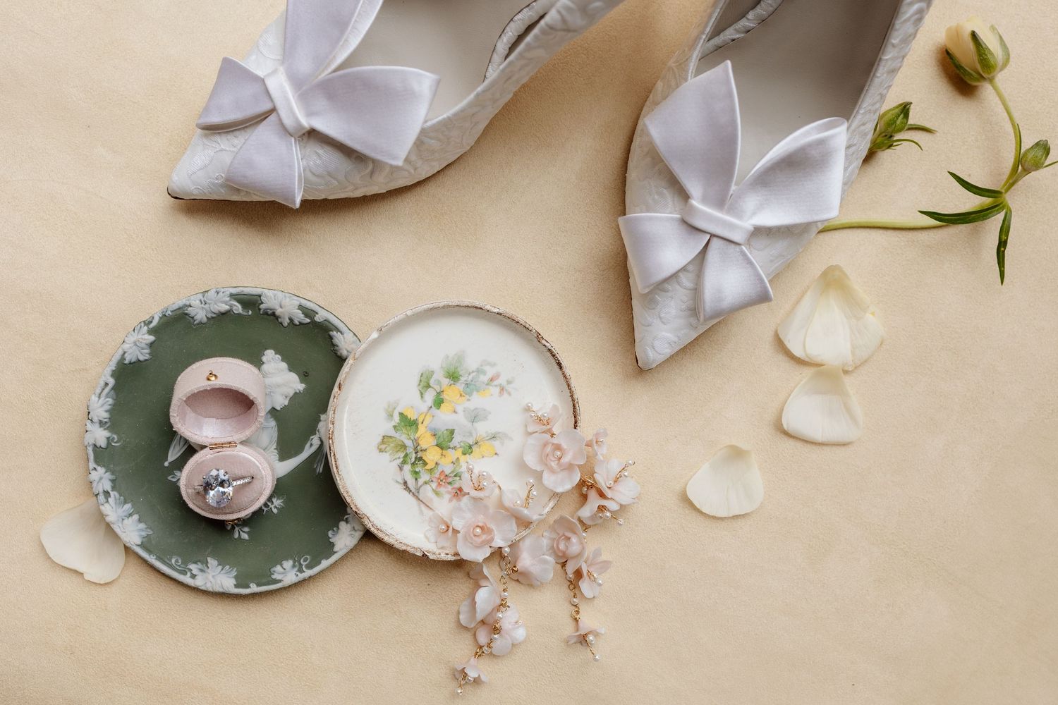 Elegant bridal accessories including white bow shoes, small plates with rings, and delicate floral elements on beige background.