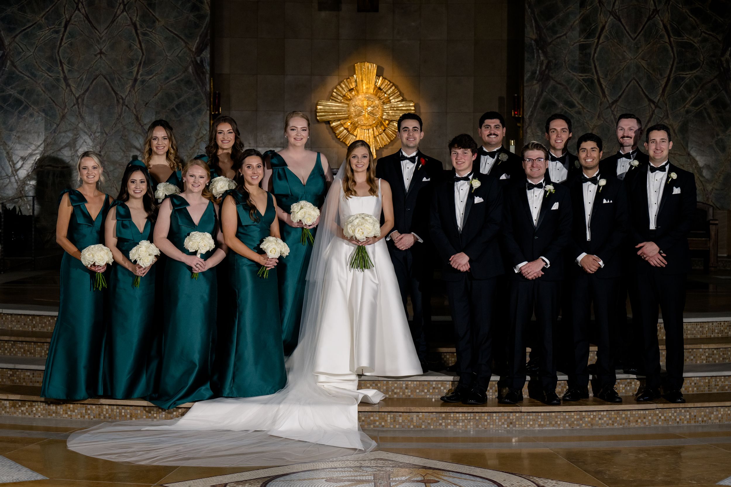 Wedding party portrait with bridesmaids in teal dresses and groomsmen in black tuxedos standing on marble steps.