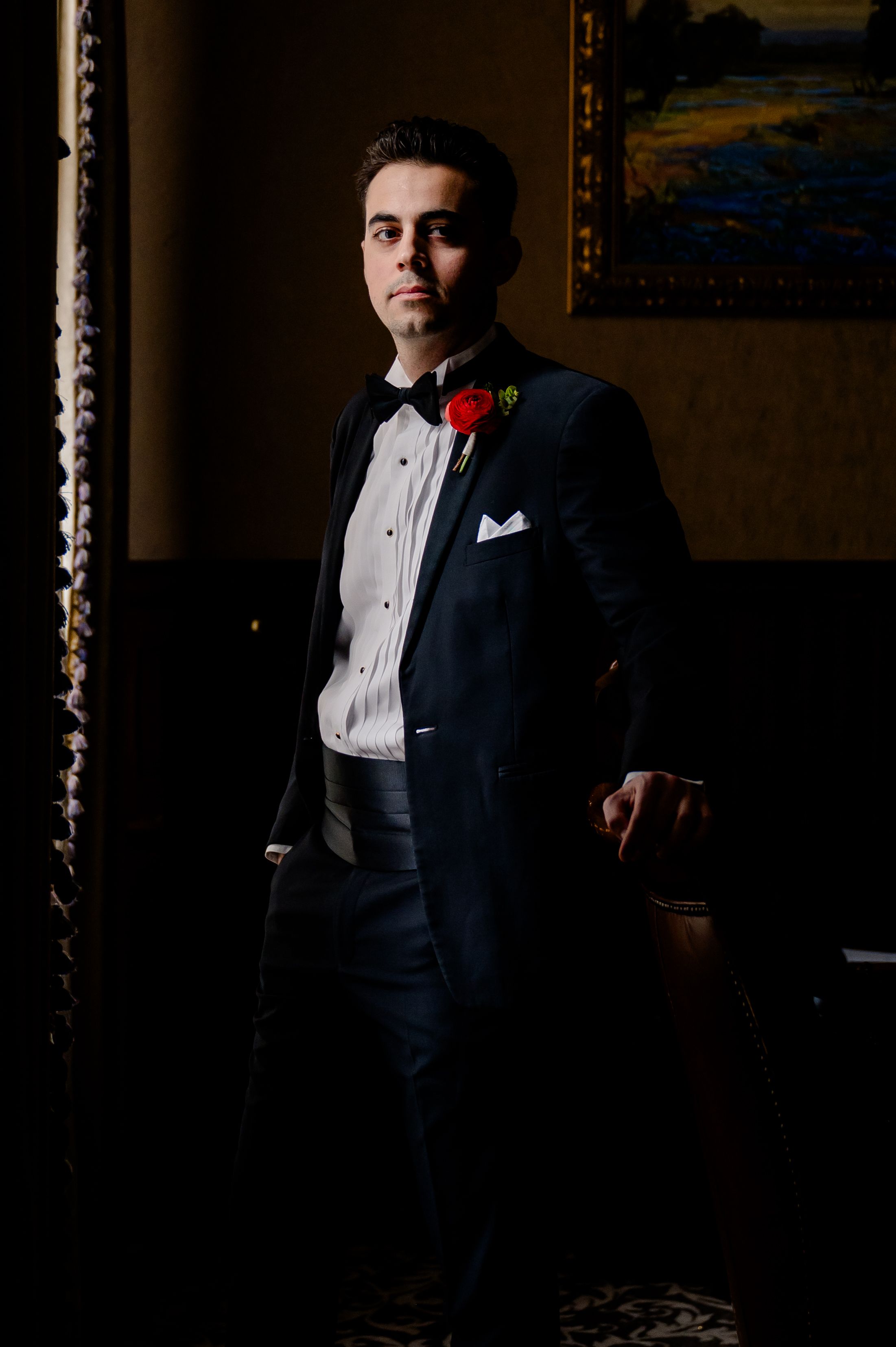 Elegant groom in classic black tuxedo with red boutonniere poses in darkly lit room with traditional decor.