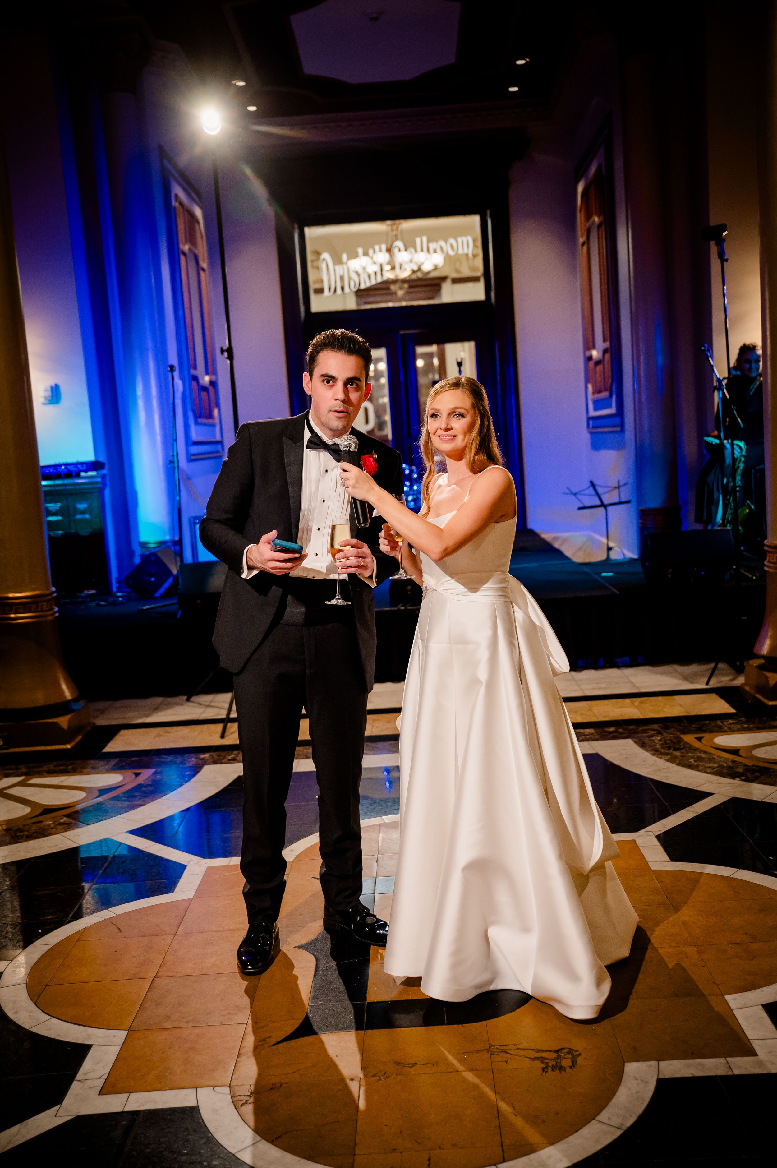 A couple in formal wedding attire raises champagne glasses for a toast under elegant lighting at an evening reception.