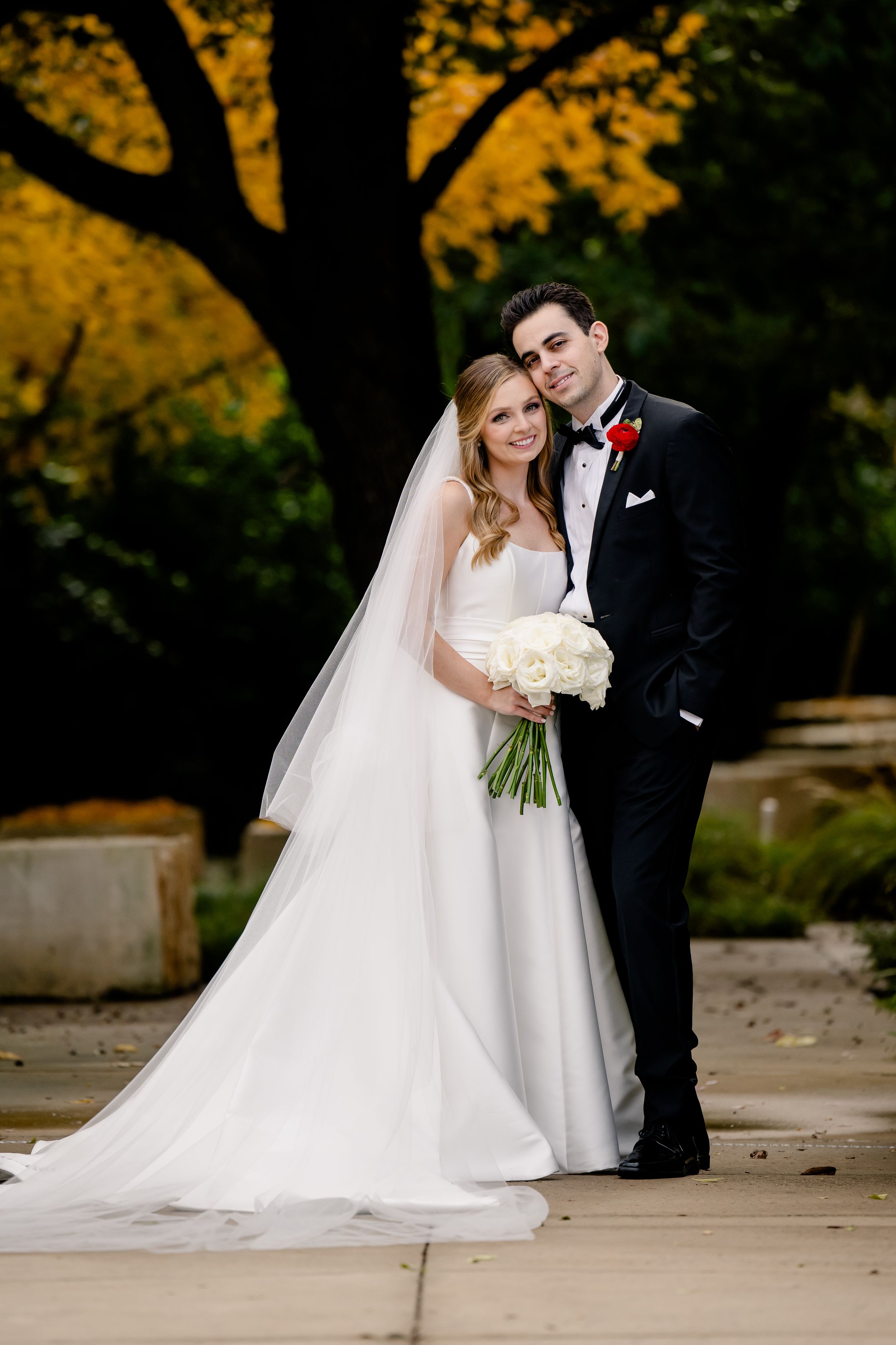A newlywed couple poses for a wedding photo against a backdrop of autumn trees with golden foliage.