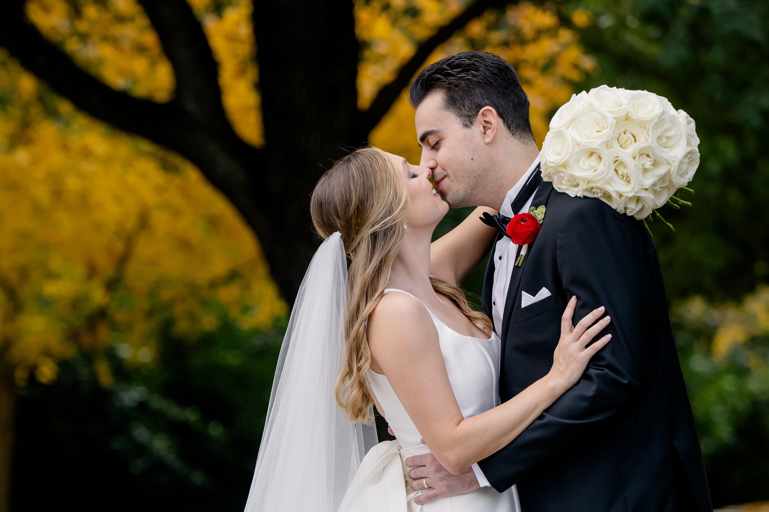 Wedding couple share romantic autumn kisses surrounded by vibrant yellow fall foliage in garden setting.