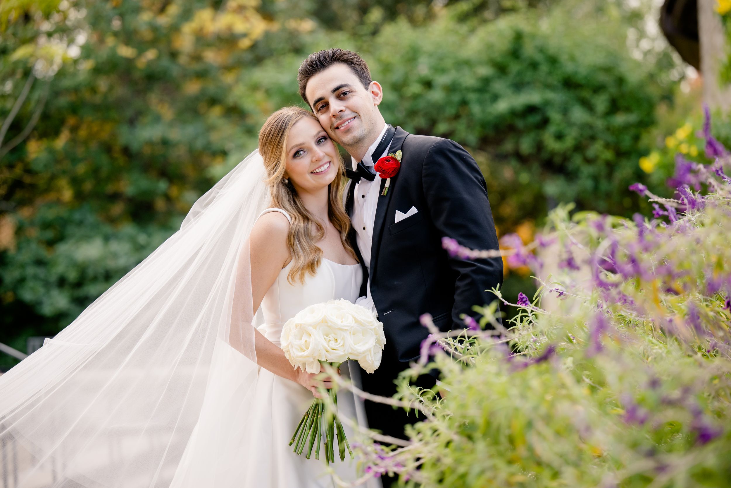 A bride and groom pose together in a garden setting with purple flowers and greenery, the bride holding a white bouquet.