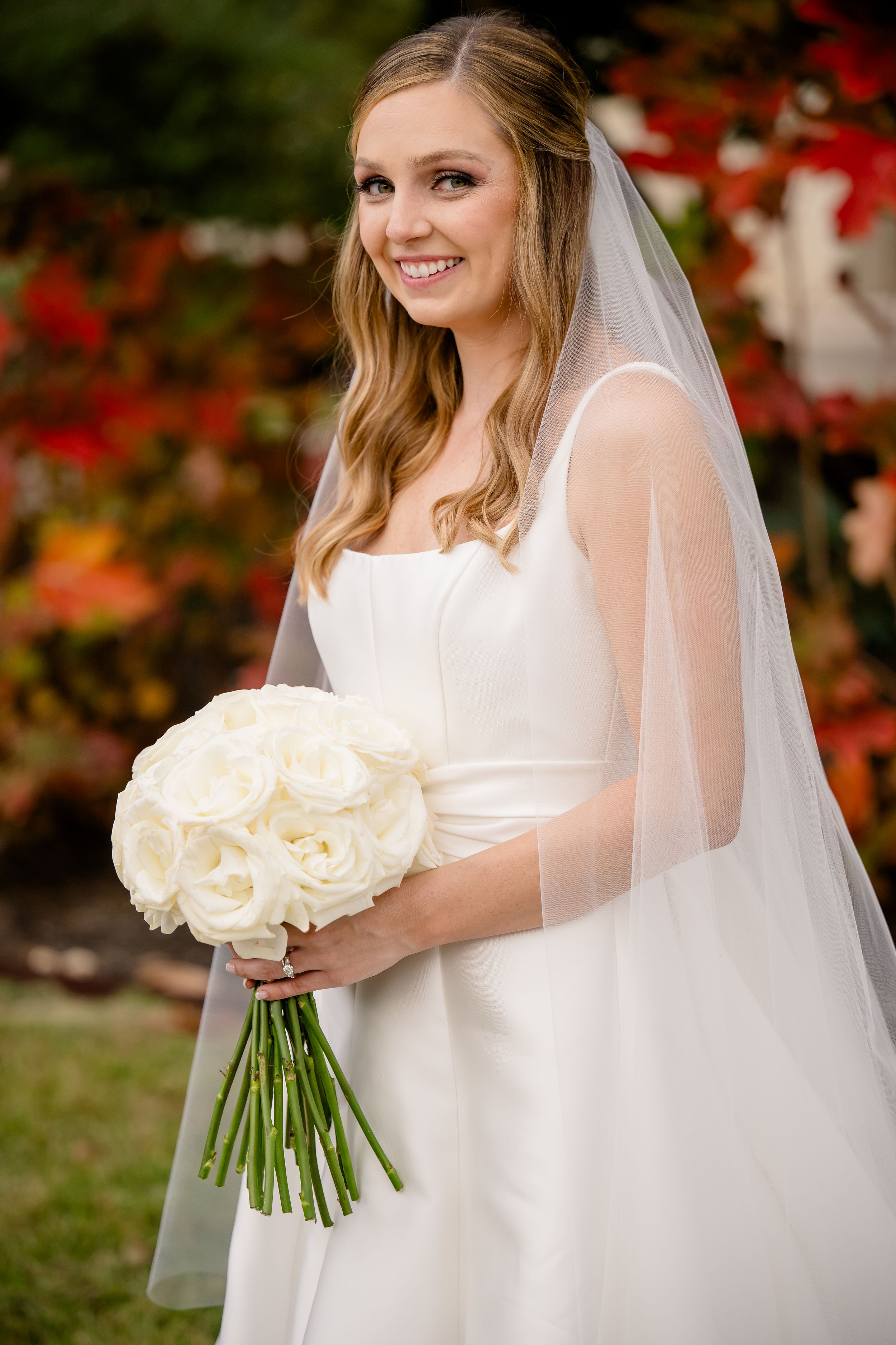 A bride in a white wedding gown holds a lush bouquet of cream roses against an autumn backdrop with red foliage.