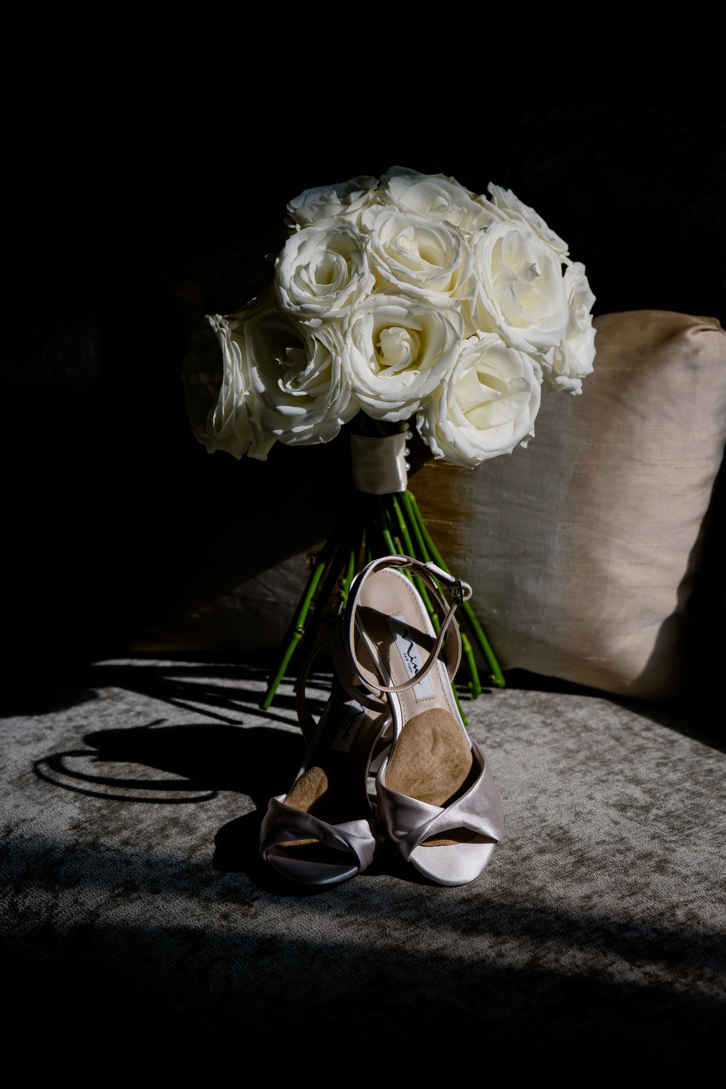 A white rose bouquet rests beside elegant strappy wedding shoes against a dark backdrop with dramatic lighting.