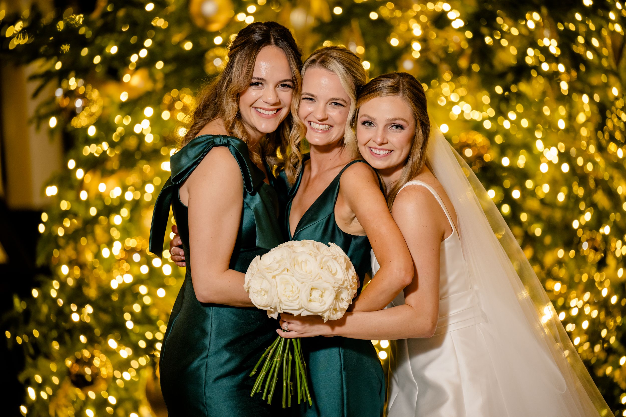 Three people in formal attire pose with a white bouquet against a glittering gold holiday light backdrop.
