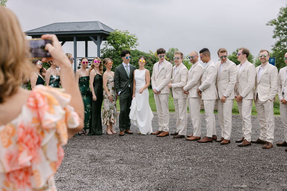 A cheerful bridal party poses in a line wearing blush and white dresses against a rustic outdoor setting.