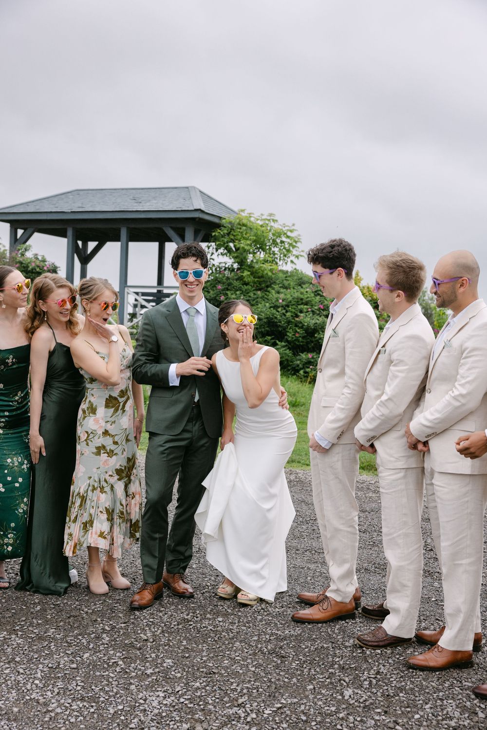 Wedding party with bridesmaids in dark dresses and groomsmen in light suits gathered outside a gazebo on overcast day.