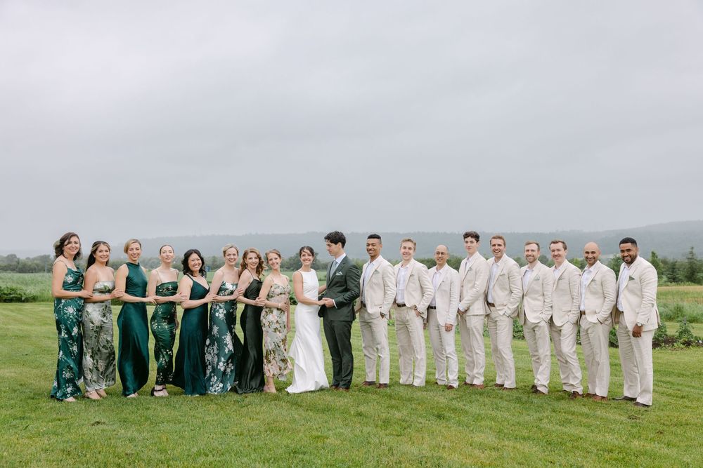 A wedding party wearing emerald green and beige formal attire poses on a grassy hilltop overlooking misty countryside.