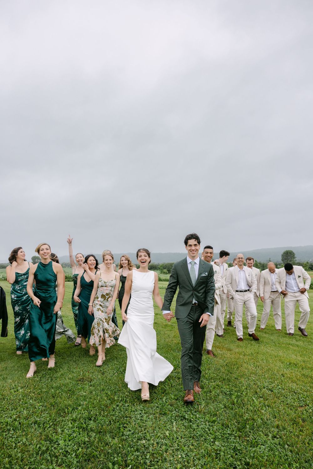 Wedding party walking together across a green field on an overcast day with bridesmaids in emerald dresses.