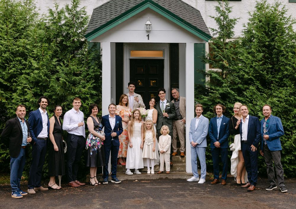 Group photo by a white chapel with a green roof on a wooded property during a special occasion.